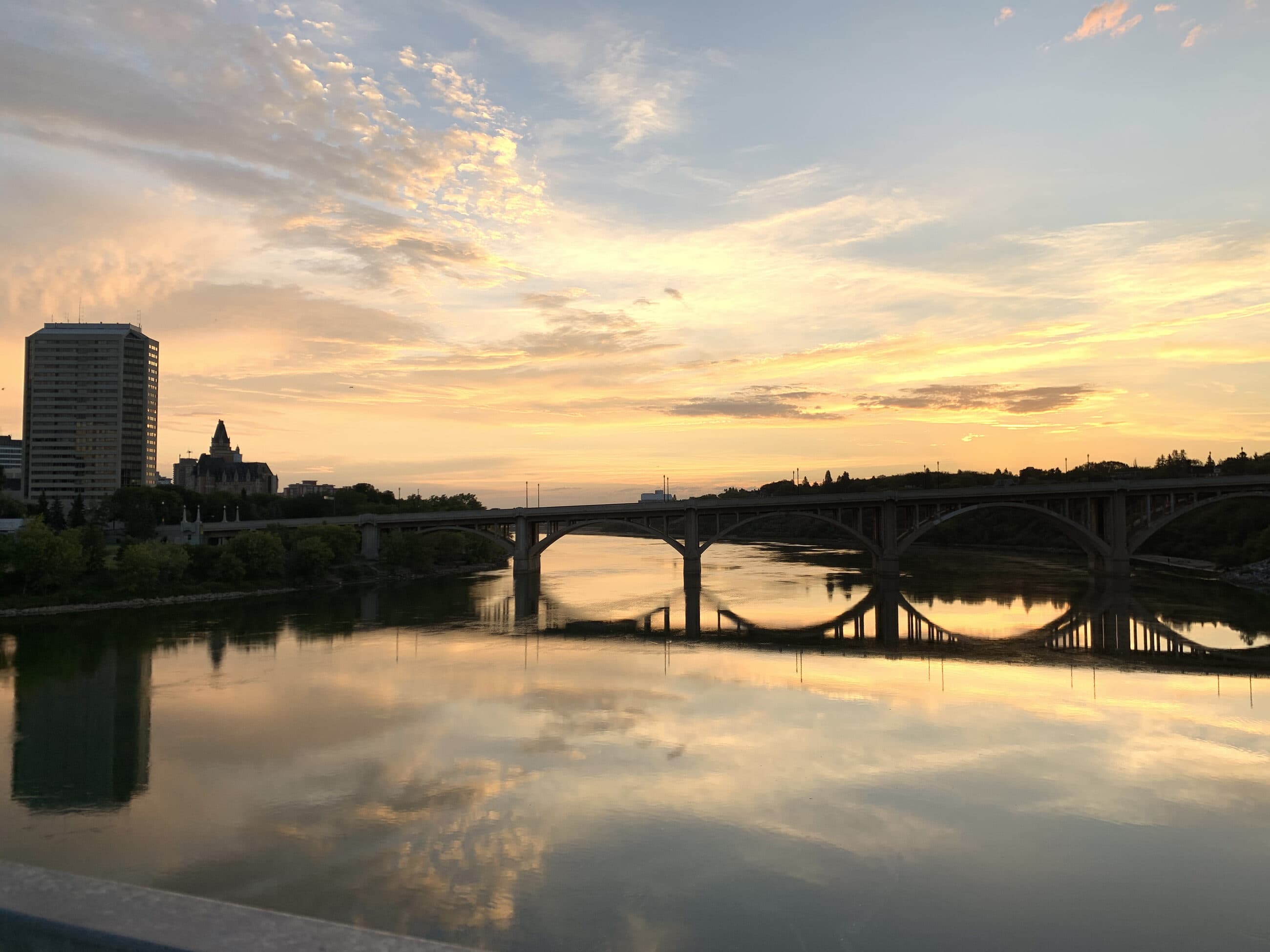The Broadway Bridge mirrored in the South Saskatchewan River under a gold sunrise sky
